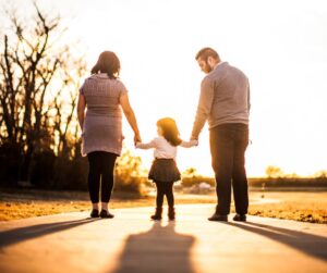 mother, father, and little girl standing in the sunlight on a side walk with backs to camera