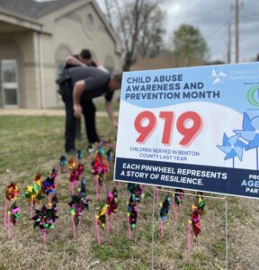 Police office putting pinwheels in the ground behind a sigh stating "919 Children Served in 2023 at the Children & Family Advocacy Center"