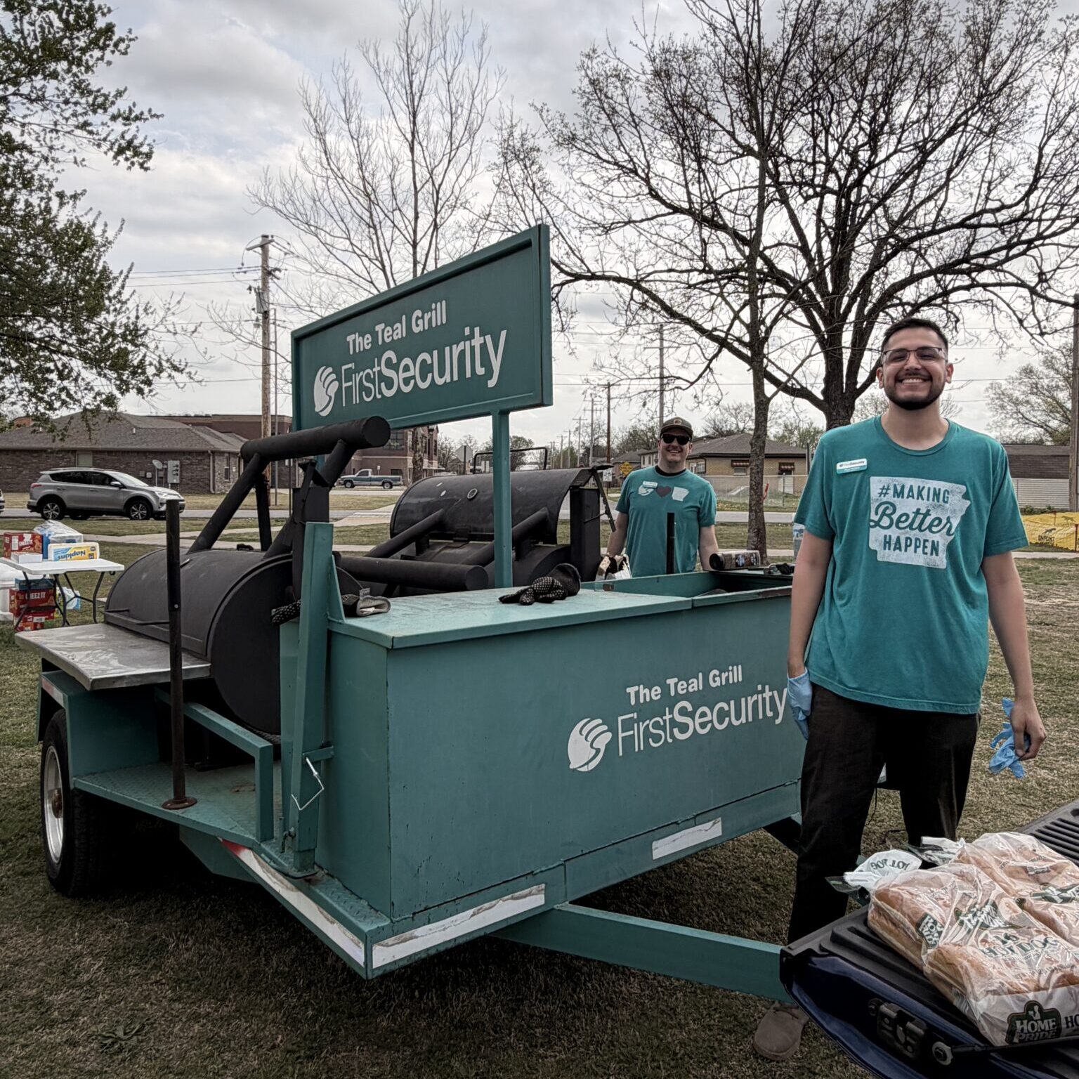 olunteers stand beside the First Security Teal Grill food trailer at an outdoor community event, preparing food for attendees.