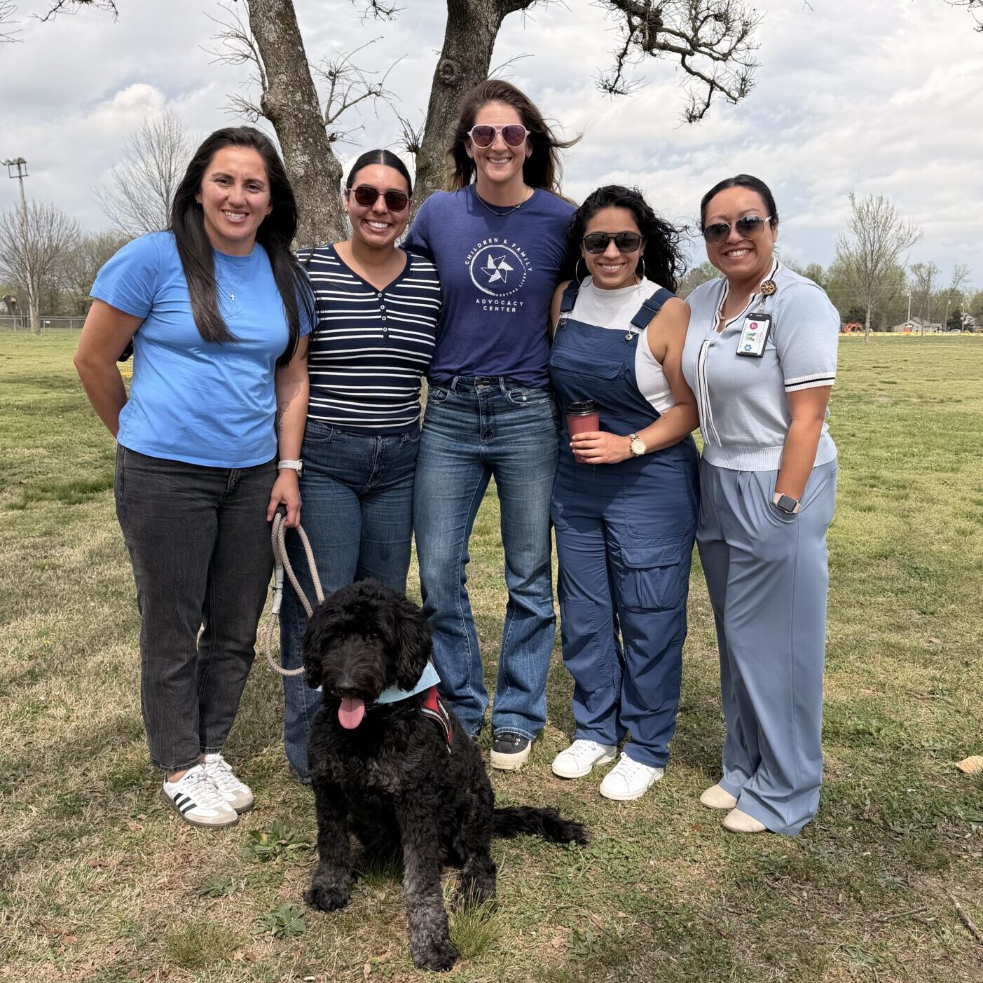 Five community partners pose with a therapy dog under a large tree at a Child Abuse Prevention Month event held outdoors.