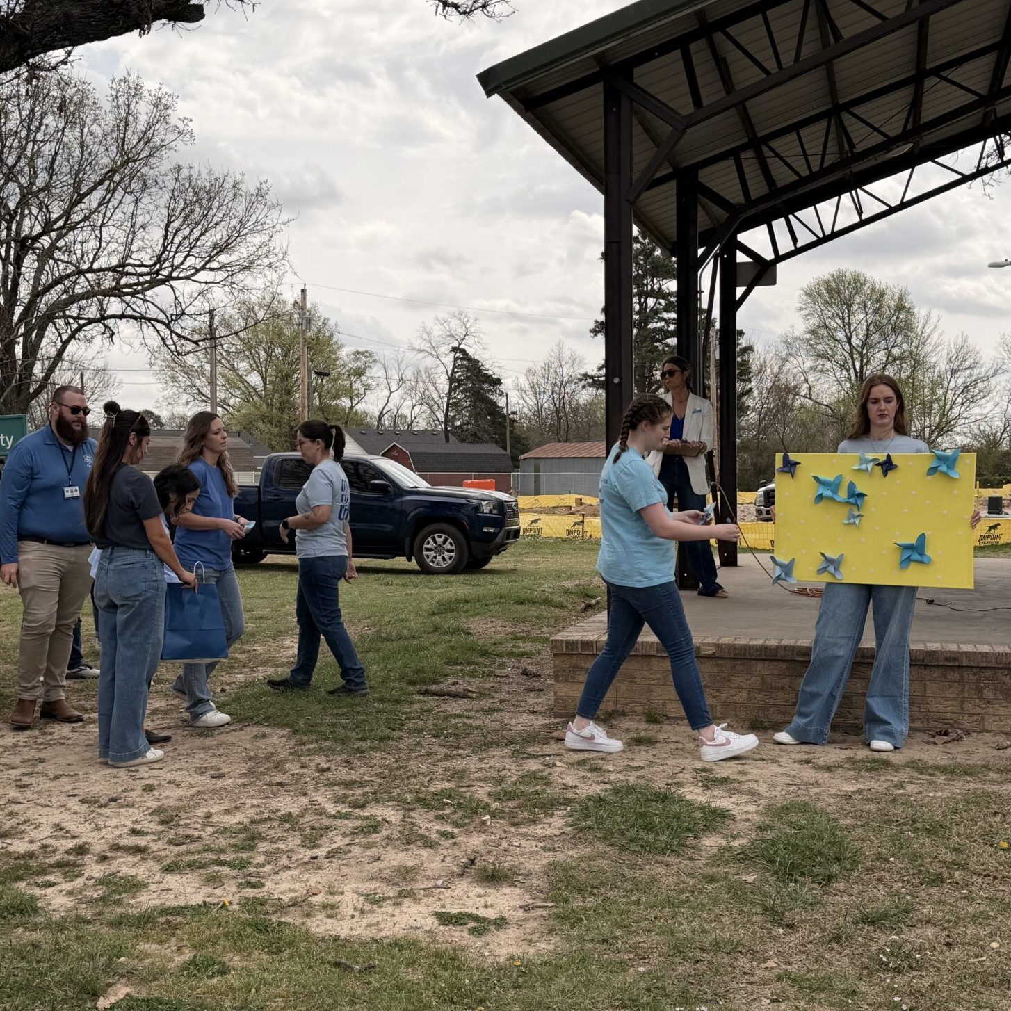 Participants gather near a pavilion stage during a community event, with one person carrying a yellow display board decorated with blue pinwheels.