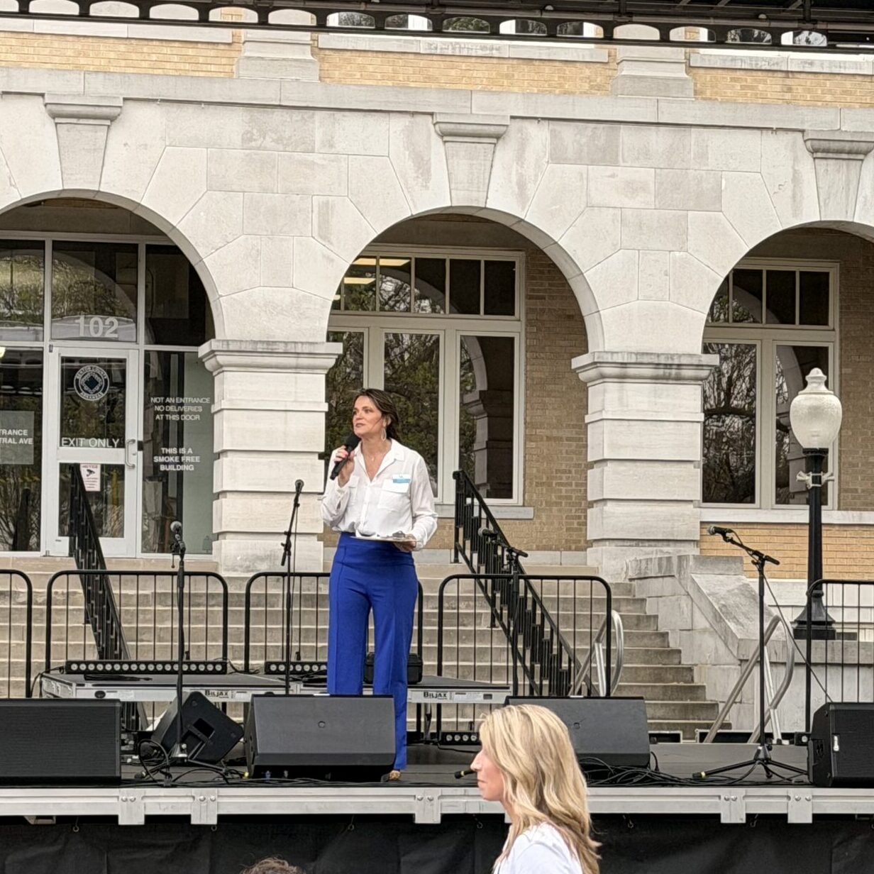 Speaker addresses attendees from an outdoor stage during a community awareness event while audience members listen from the plaza below.