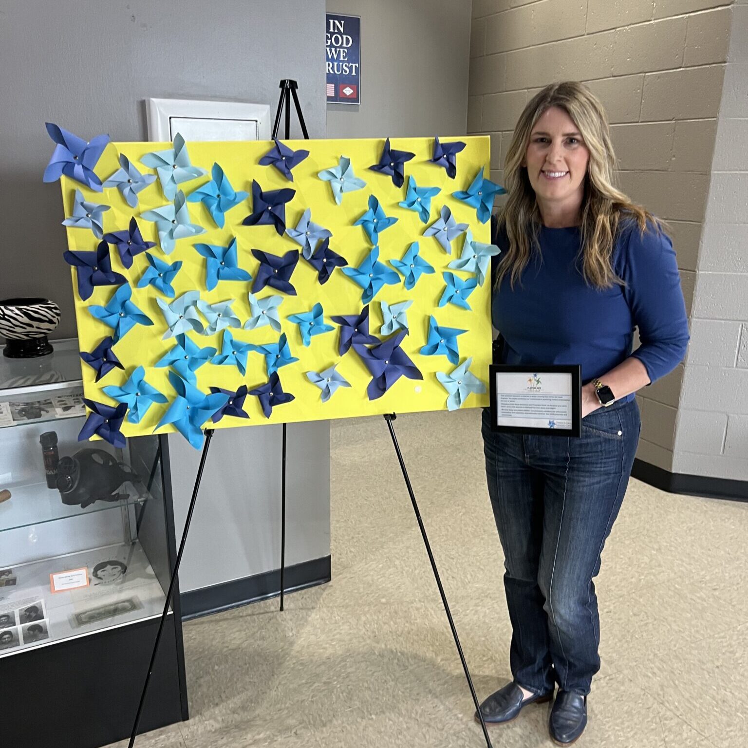 Person stands beside a yellow display board decorated with blue pinwheels while holding a framed certificate during a child abuse prevention awareness event.