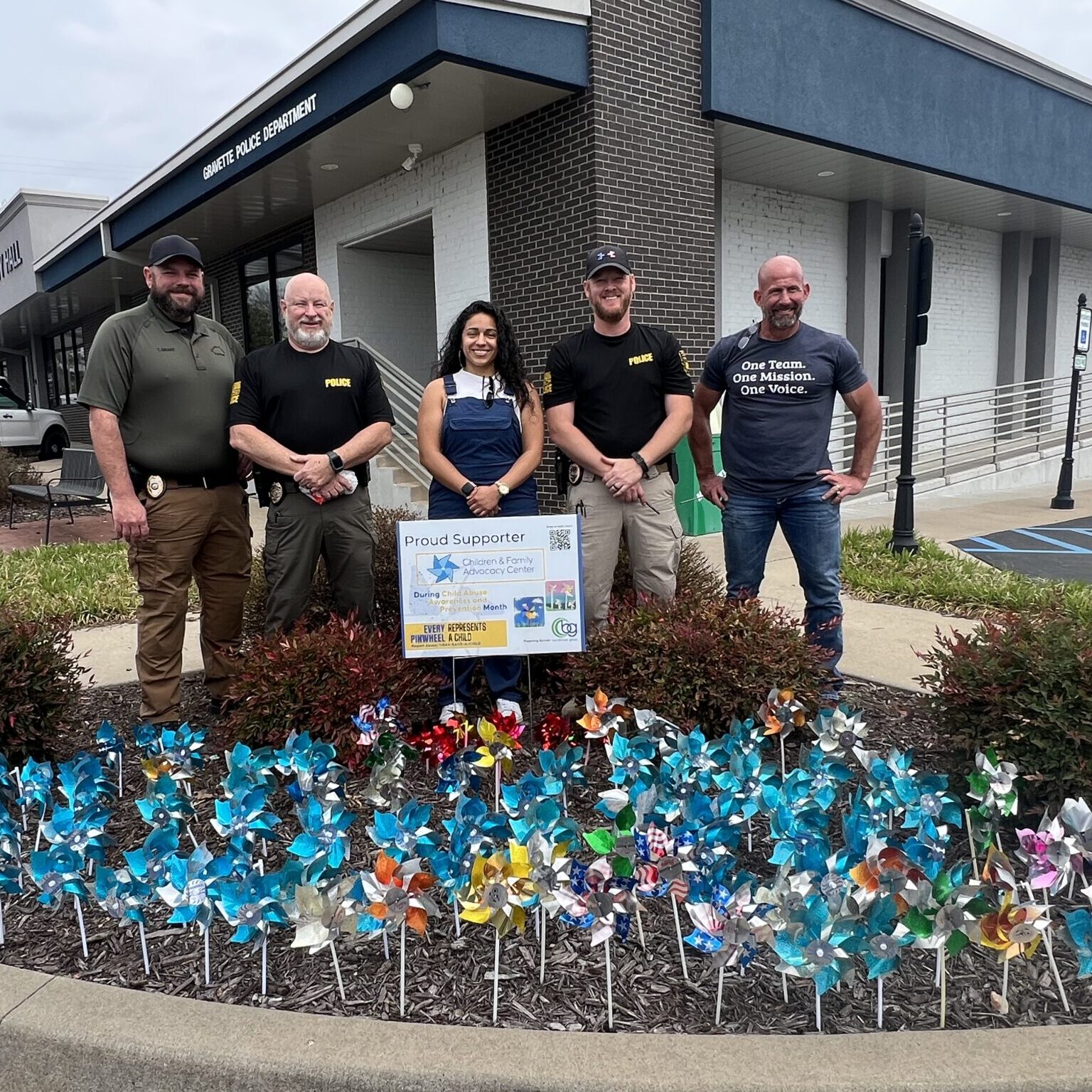 Community members pose in front of a police department building and a pinwheel garden in support of Child Abuse Prevention Month.
